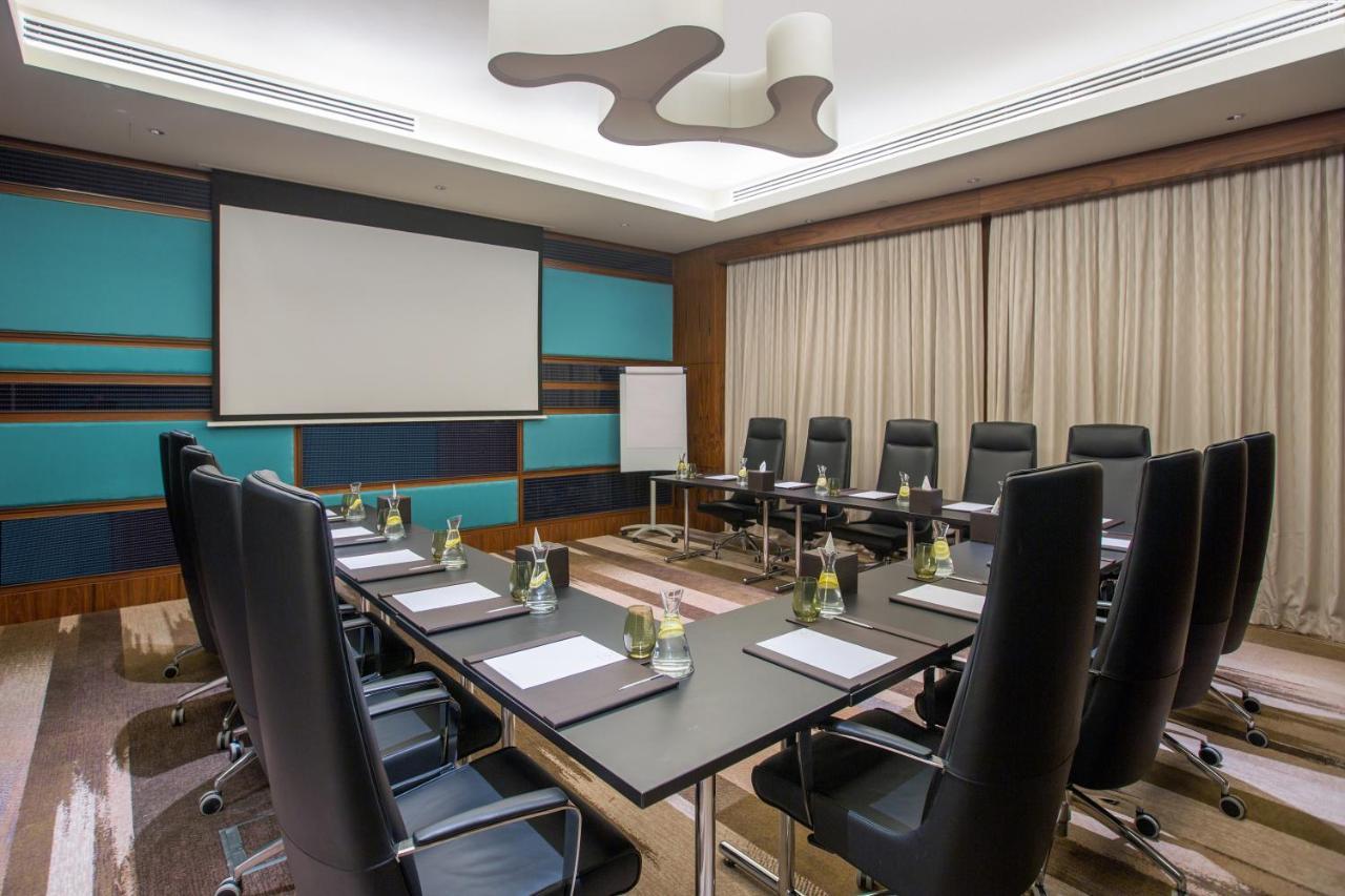 Hotel Millennium Plaza Doha Exterior foto The photo shows a modern conference room. It features a long table with several black leather chairs arranged around it. On the table, there are notepads and pens for each seat. A projector screen is visible on one wall, and a flip chart stand is pla