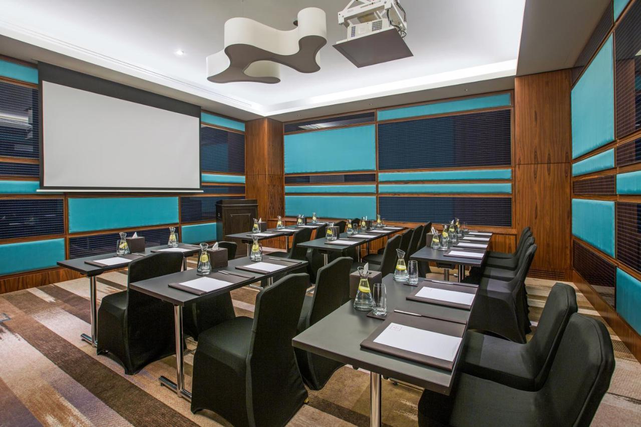 Hotel Millennium Plaza Doha Exterior foto The photo shows a modern conference room. It features several rectangular tables arranged in a classroom-style setup, each with a black chair. On each table, there are notepads and pens, along with bottles of water. The walls are accented with a mix 