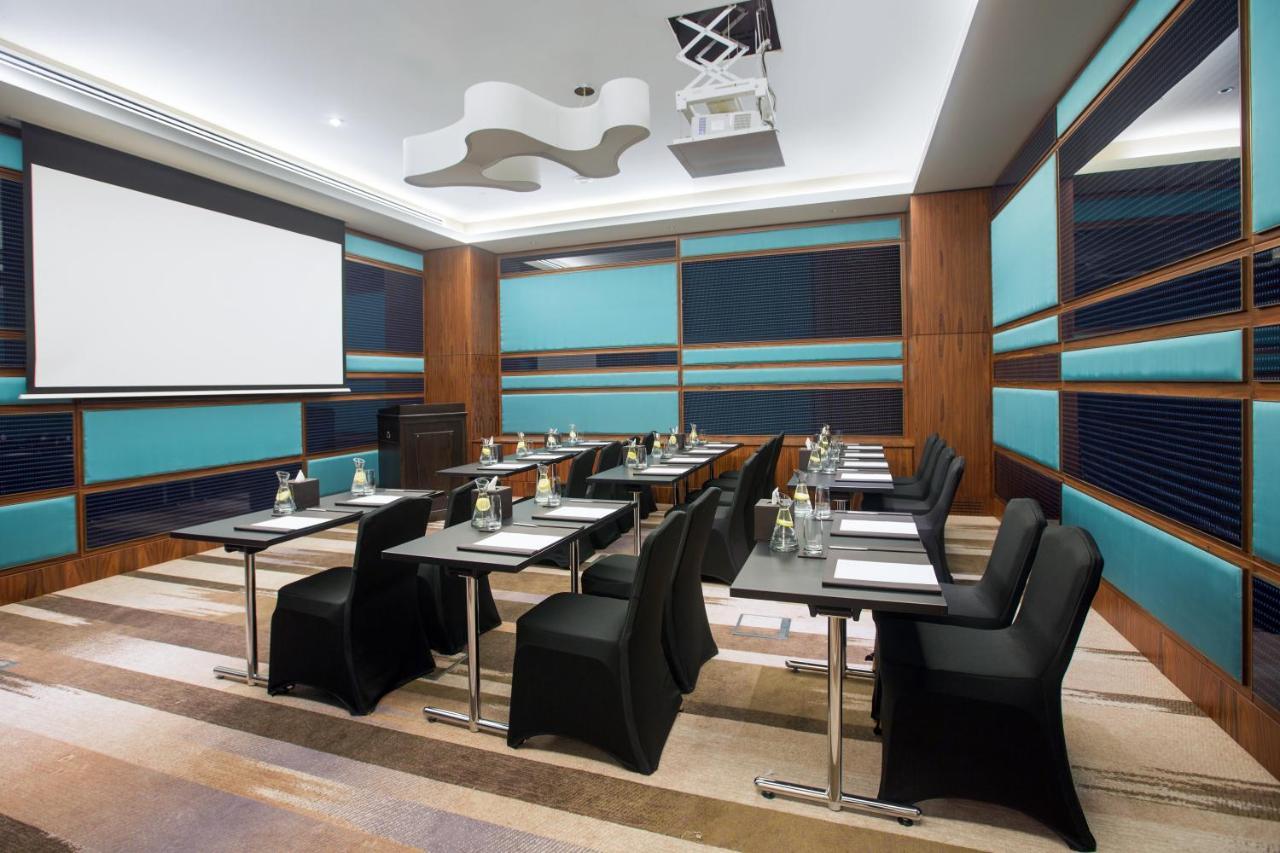 Hotel Millennium Plaza Doha Exterior foto The photo shows a modern conference room or meeting space. It features several black chairs arranged around tables, with each table set up with notepads and water bottles. The walls have a combination of wooden panels and blue accents. A large screen