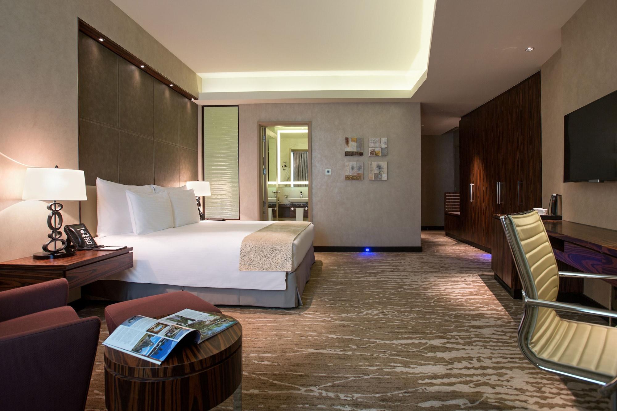Hotel Millennium Plaza Doha Exterior foto The photo shows a modern hotel room featuring a spacious layout. There's a large bed with crisp white linens centered in the room. To the left, there is a small table with a magazine on top, and a sleek chair nearby. The decor is contemporary, with s