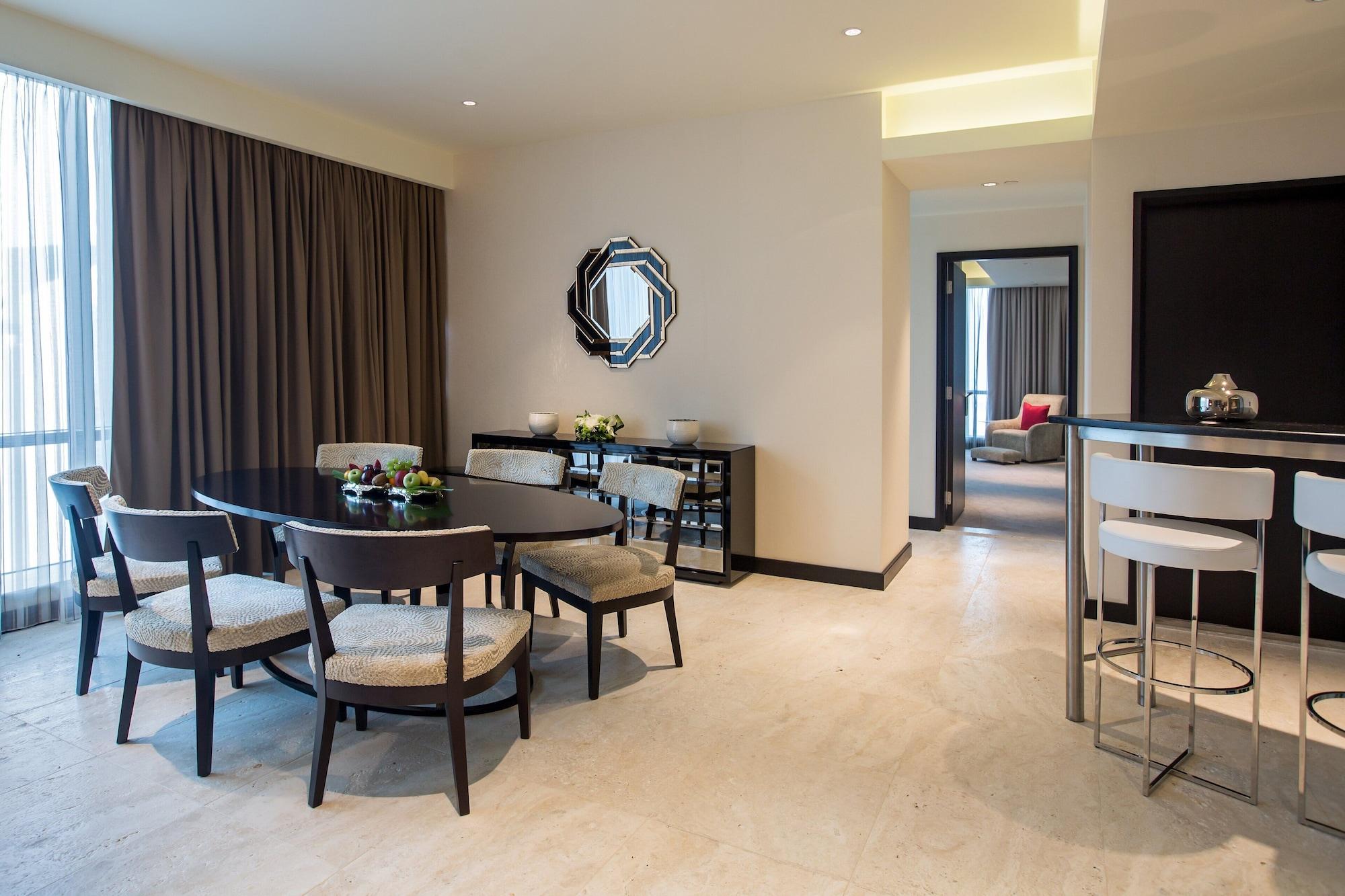Hotel Millennium Plaza Doha Exterior foto The photo shows an elegant living space. On the left, there is a dining table surrounded by several chairs. The table appears to have a centerpiece with some decorative items. In the background, large windows are covered by long, neutral-colored curt