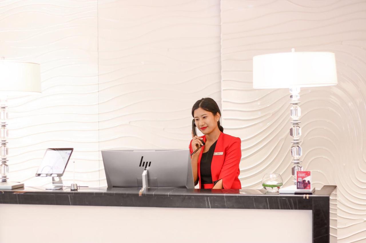 Hotel Millennium Plaza Doha Exterior foto The image shows a receptionist at a hotel or office front desk. The receptionist, dressed in a red blazer, is seated behind a sleek desk with a modern computer monitor. She is speaking on the phone and appears to be engaged in her work. The backgroun