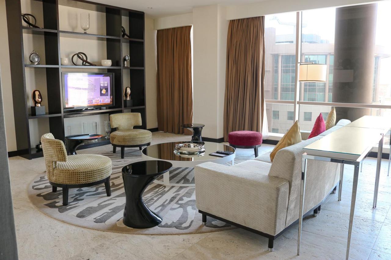 Hotel Millennium Plaza Doha Exterior foto The photo shows a modern living room interior. There are several stylish seating arrangements, including armchairs and a sofa. A round coffee table is placed in the center, surrounded by decorative items. Behind the seating area, there is a large win