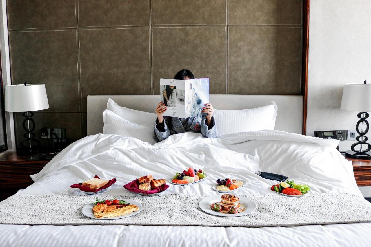 Hotel Millennium Plaza Doha Exterior foto The photo depicts a person sitting in a bed, covered with white bedding. The individual is holding a magazine in front of their face, suggesting they are either reading or looking at something interesting. Around them on the bed are several plates of