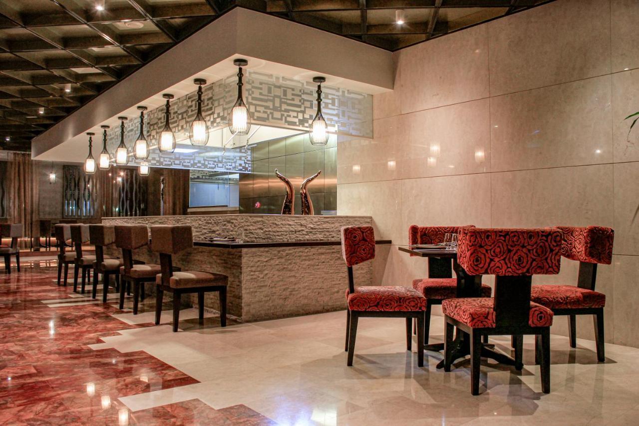 Hotel Millennium Plaza Doha Exterior foto The photo depicts a modern restaurant interior. It features a bar area with a stone counter and stylish lighting fixtures hanging overhead. The seating consists of several dark wooden chairs with red-patterned upholstery arranged around small tables.