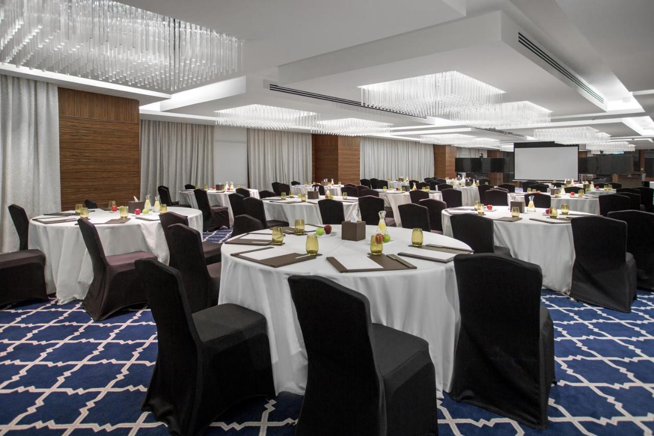 Hotel Millennium Plaza Doha Exterior foto The photo shows a spacious banquet or conference hall set up for an event. The room features round tables covered with white tablecloths, each adorned with utensils and small decorative items. The chairs are primarily black, and the flooring has a bl