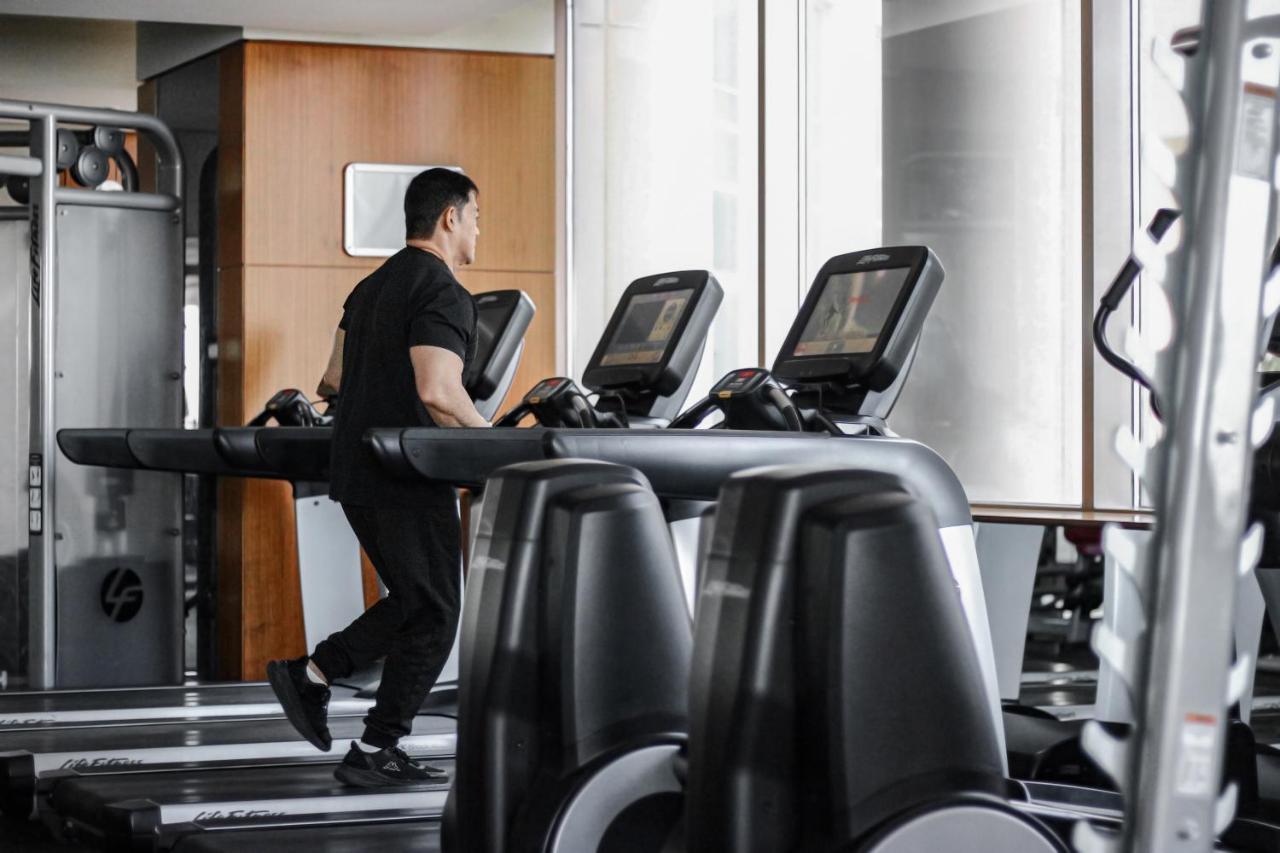 Hotel Millennium Plaza Doha Exterior foto The photo shows a man exercising on a treadmill in a gym setting. He is wearing a black workout outfit and appears to be focused on his exercise. The background features large windows letting in natural light, and there are other fitness machines vis