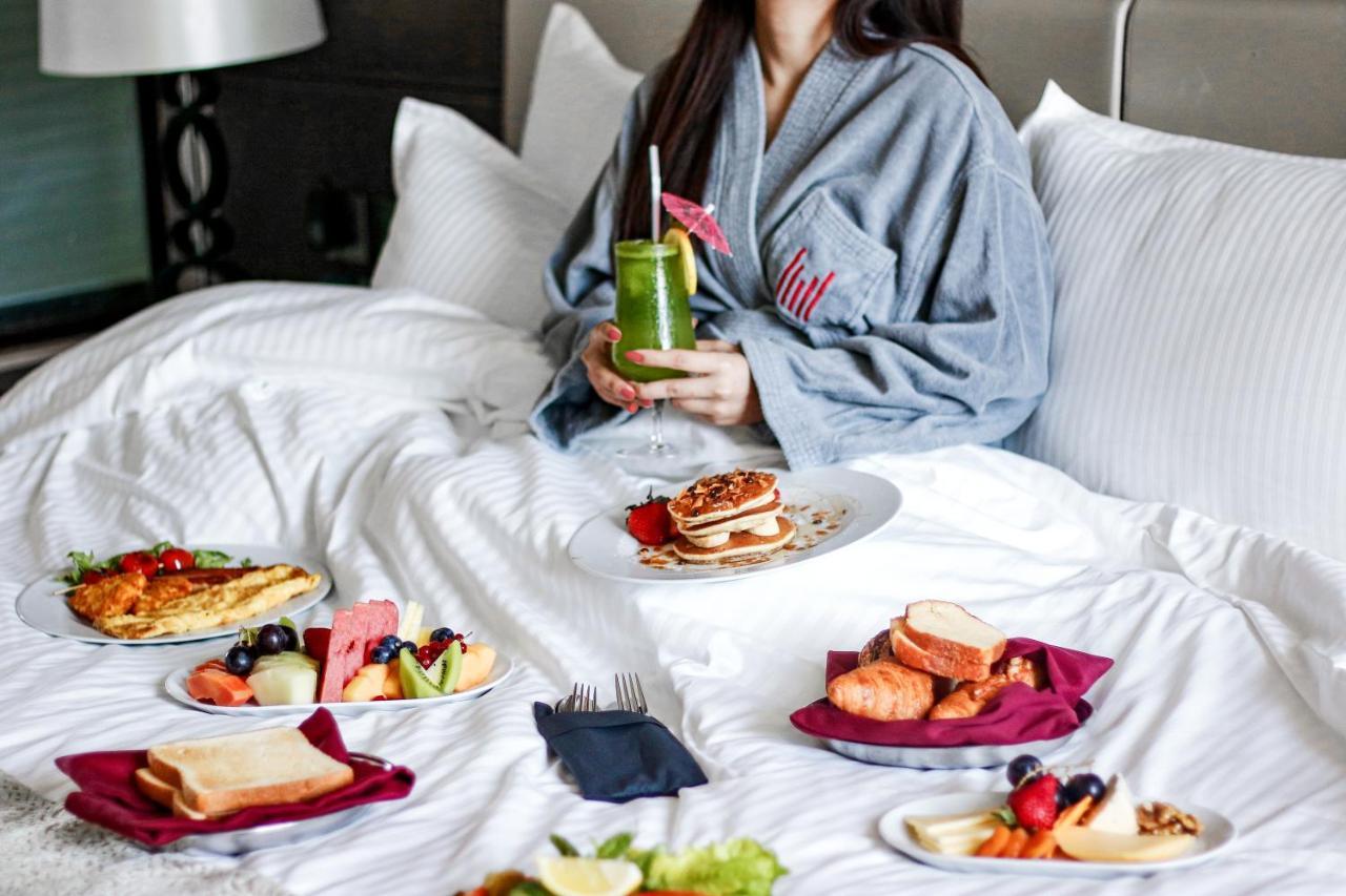 Hotel Millennium Plaza Doha Exterior foto The image shows a person sitting in bed, wearing a gray robe. Surrounding the person are several plates of breakfast food. There is a variety of items, including pancakes, fruit, pastries, bread, and a drink that appears to be a green smoothie or jui