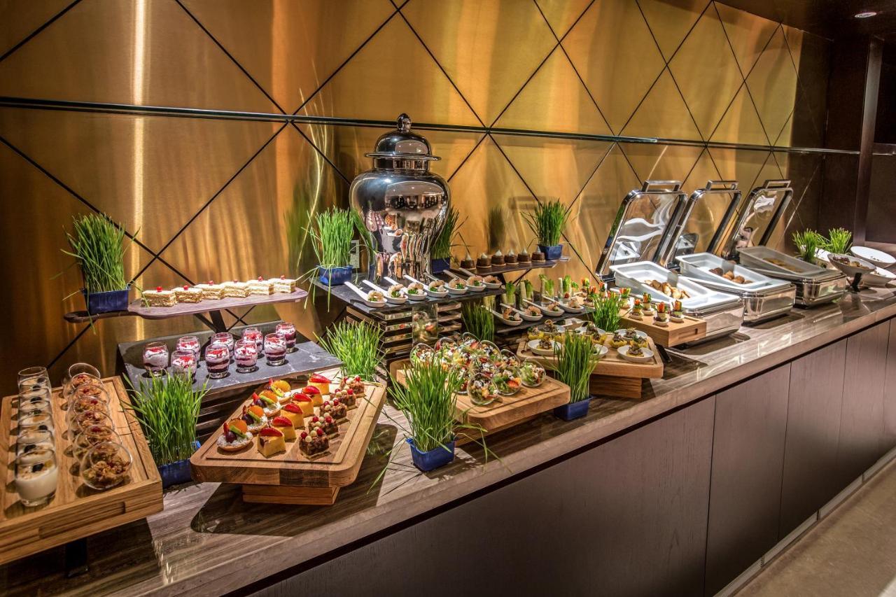 Hotel Millennium Plaza Doha Exterior foto The photo displays an elegantly arranged buffet setup featuring a variety of delicacies. There are multiple plates of assorted appetizers and desserts on wooden trays, including small portions that look gourmet. In the background, a large silver beve