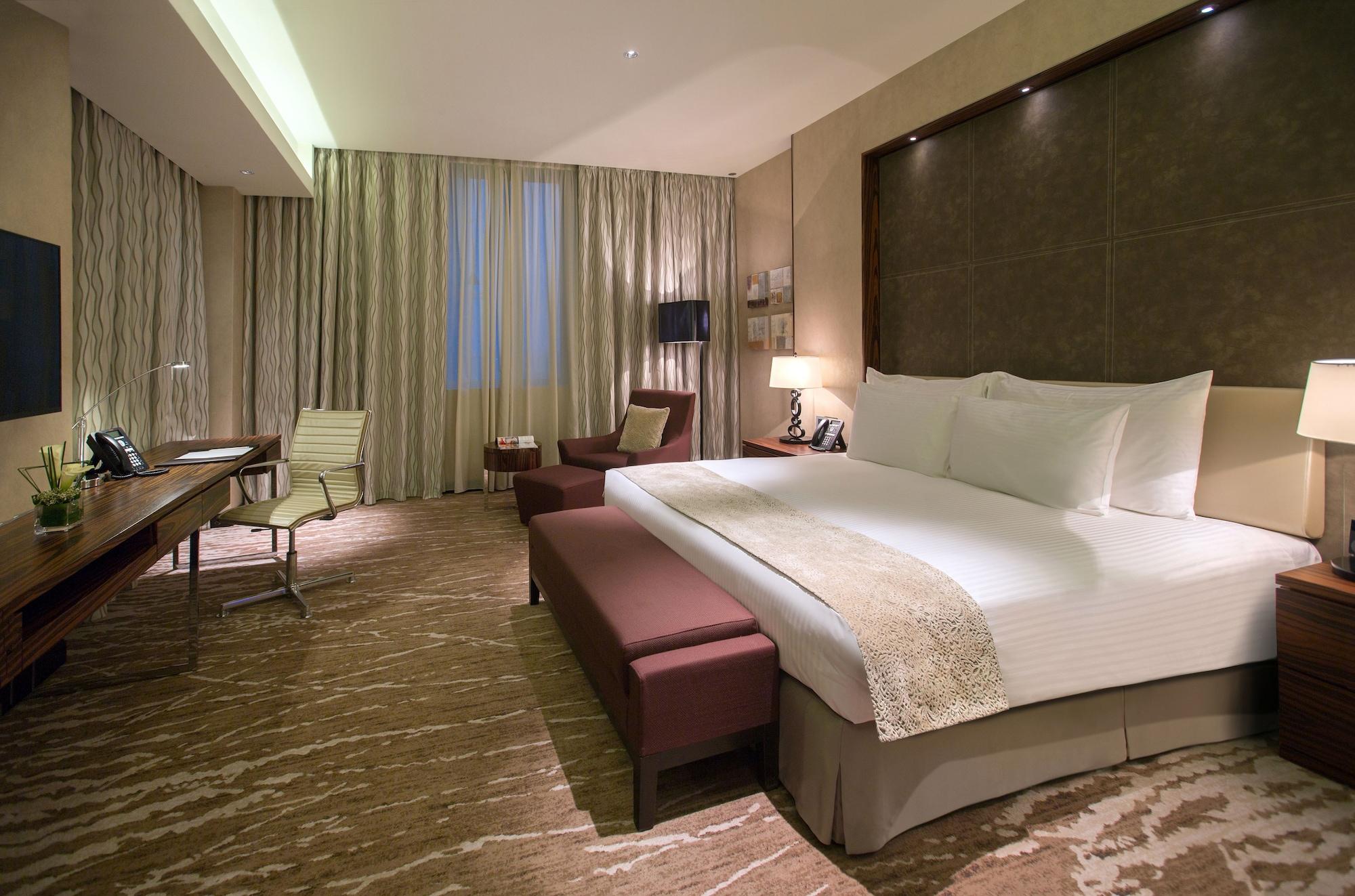 Hotel Millennium Plaza Doha Exterior foto The photo shows a modern hotel room featuring a comfortable king-sized bed with white linens and decorative pillows. There is a cozy seating area with a chair and a small couch or bench at the foot of the bed. A stylish desk with a chair is positione