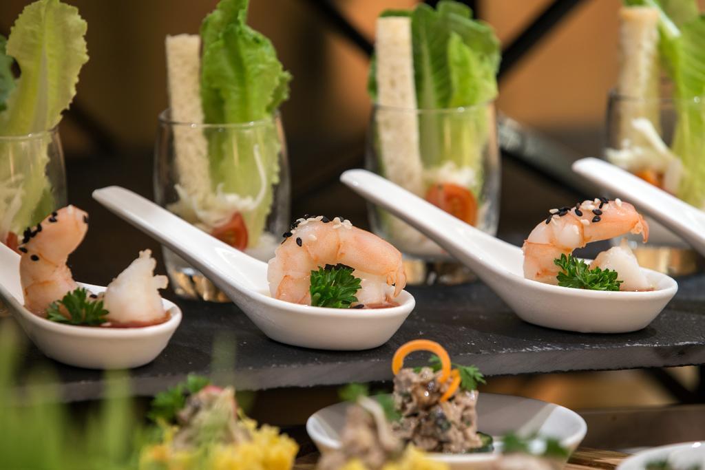 Hotel Millennium Plaza Doha Exterior foto The photo showcases a visually appealing arrangement of gourmet dishes. In the foreground, there are small white spoons holding shrimp, garnished with a sprig of parsley. Behind them, there are small glass cups containing layers of ingredients, likel