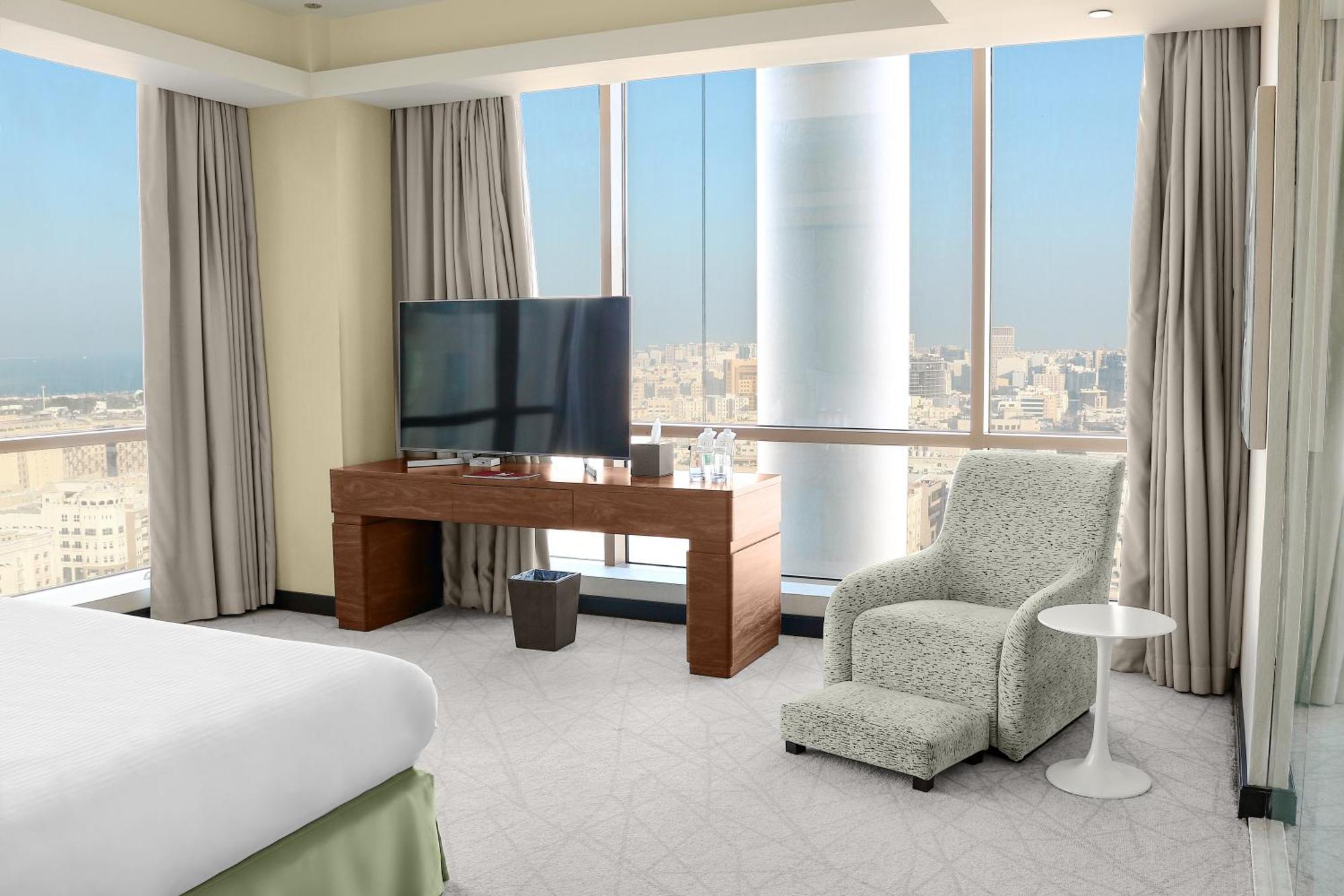 Hotel Millennium Plaza Doha Exterior foto The photo shows a modern hotel room with large windows that provide a view of the city. On the right side, there is a comfortable armchair next to a small side table. Opposite the armchair is a wooden console or desk with a flat-screen television mou