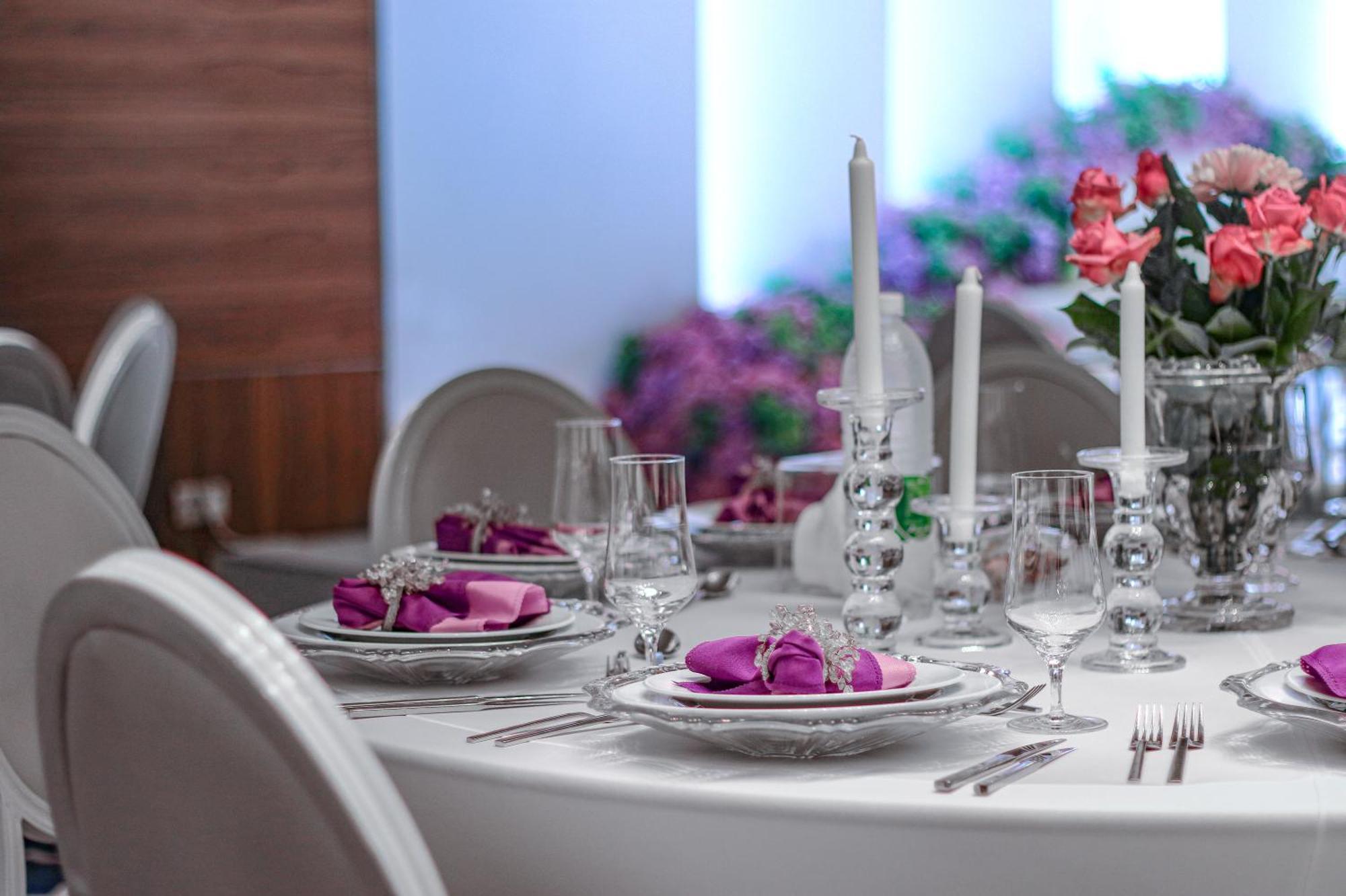 Hotel Millennium Plaza Doha Exterior foto The image shows a beautifully set dining table. The table has a white tablecloth and is arranged with elegant dinnerware, including plates with purple napkins folded decoratively. There are crystal glasses and silver utensils arranged neatly around t