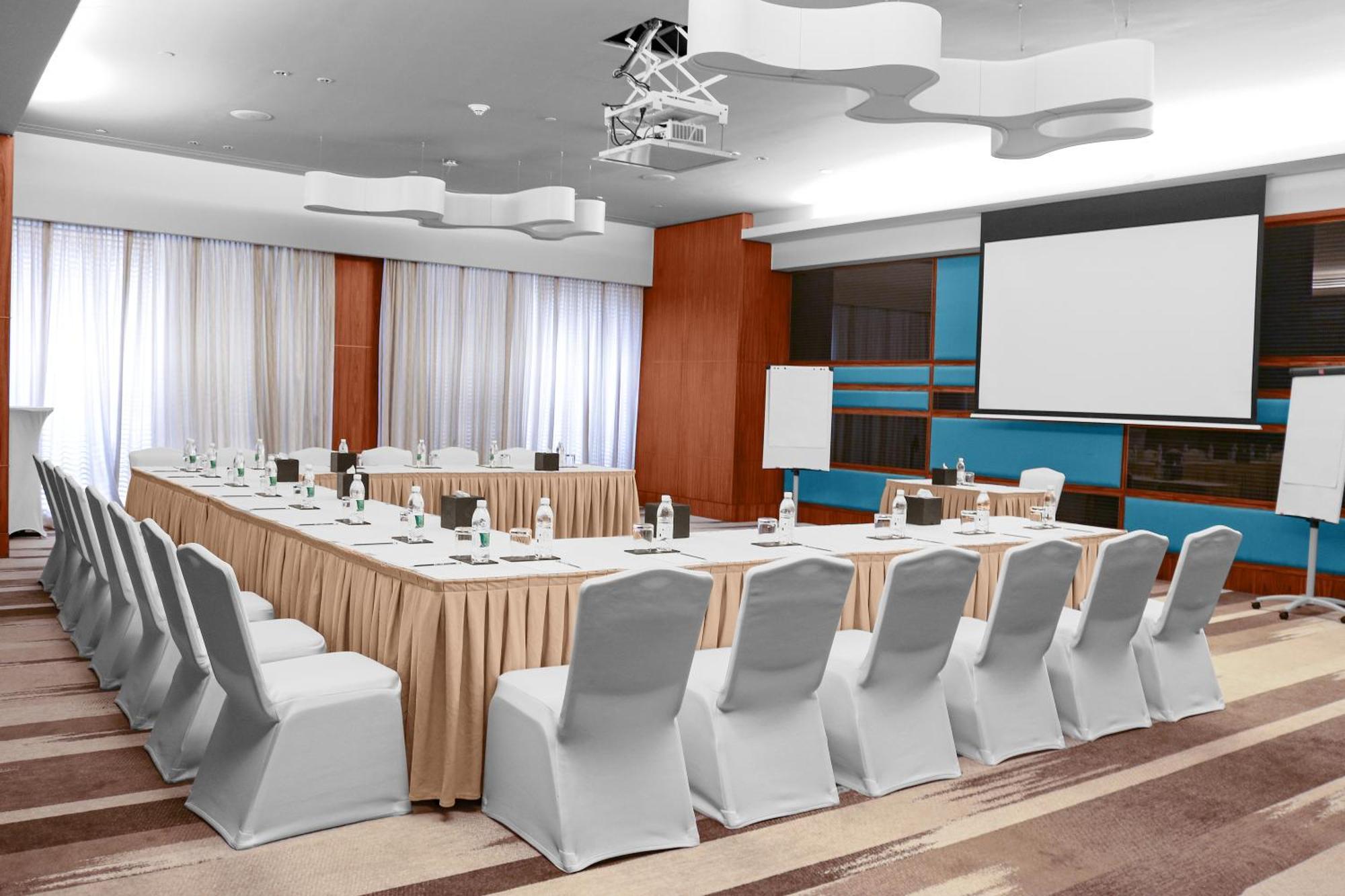 Hotel Millennium Plaza Doha Exterior foto The photo shows a conference room set up for a meeting or seminar. There is a large rectangular table in the center covered with a beige tablecloth. Around the table, there are several chairs, all covered with white chair covers. At one end of the ro