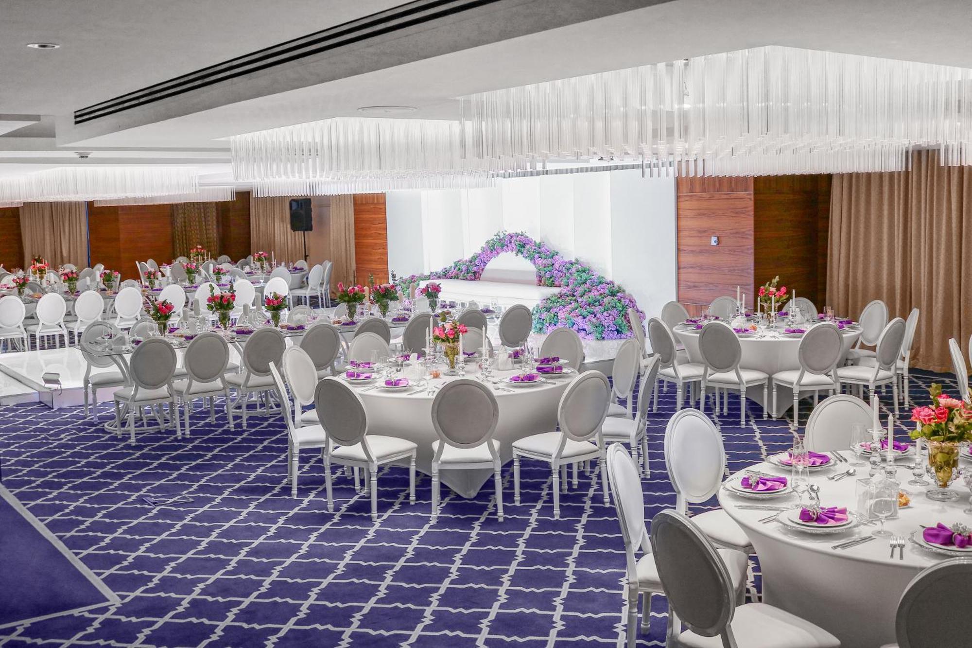 Hotel Millennium Plaza Doha Exterior foto The photo depicts an elegant banquet hall set up for an event. The room features round tables covered with white tablecloths and adorned with floral centerpieces. There are white chairs arranged around the tables, and the floor has a patterned blue c