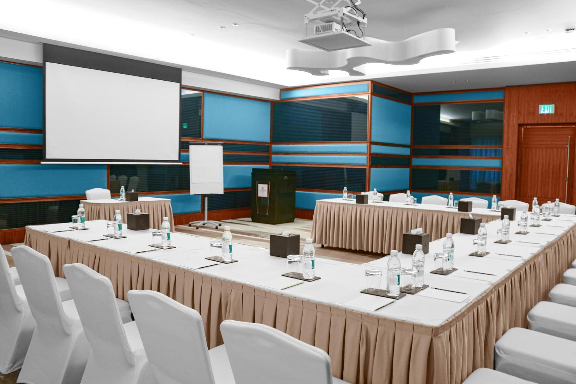 Hotel Millennium Plaza Doha Exterior foto The photo shows a conference room setup. There are multiple tables arranged in a U-shape, covered with white tablecloths and beige skirts. Each table has small notepads, pens, and water bottles placed on them. In the center, there is a larger table w