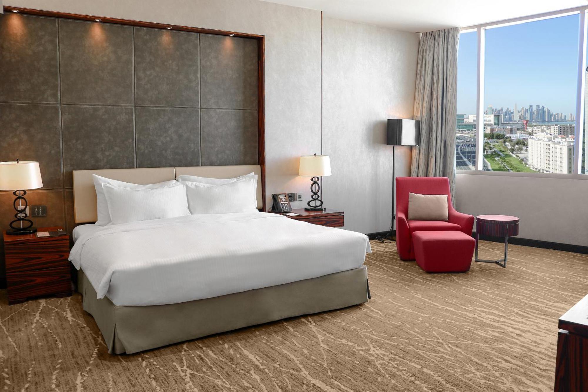 Hotel Millennium Plaza Doha Exterior foto The photo shows a modern hotel room featuring a large bed with white linens positioned against a gray accent wall. There are bedside tables with lamps on either side of the bed. To the side, there's a comfortable red armchair with a small table nearb