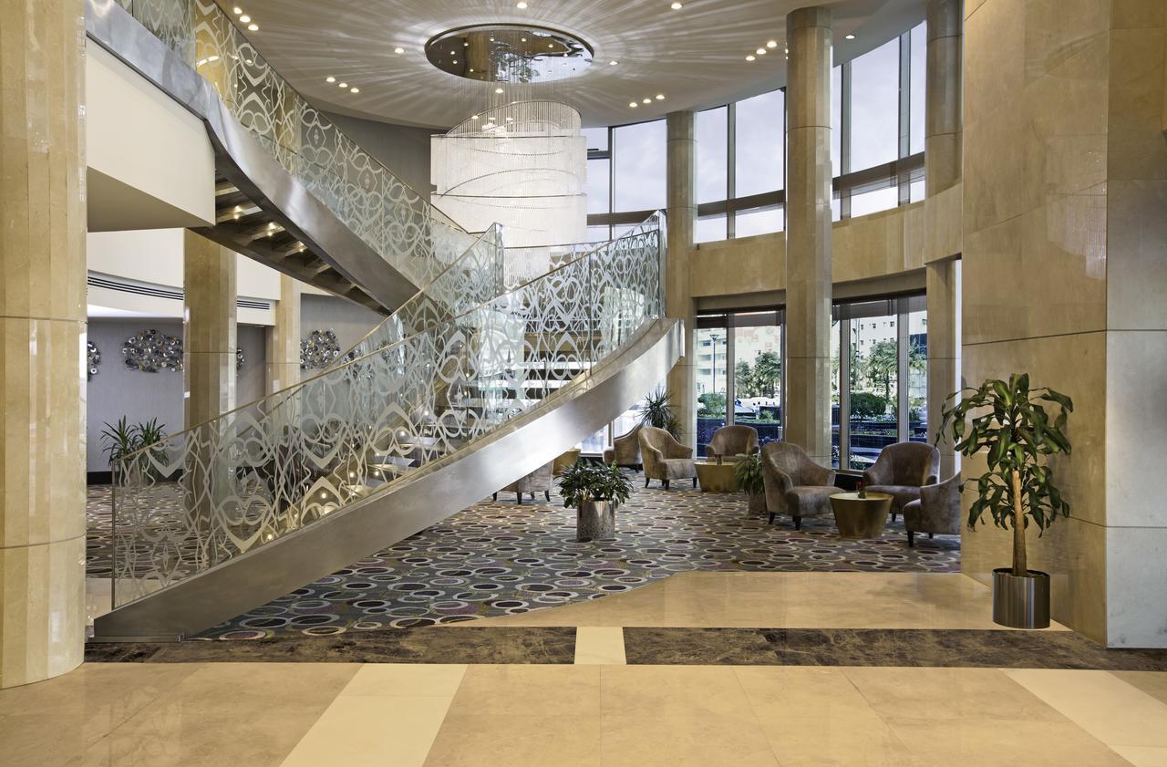 Hotel Millennium Plaza Doha Exterior foto The photo shows a modern and elegant lobby area. It features a sweeping staircase with a decorative railing that draws the eye upward. The space is filled with natural light coming through large windows, creating an inviting atmosphere. The floor has