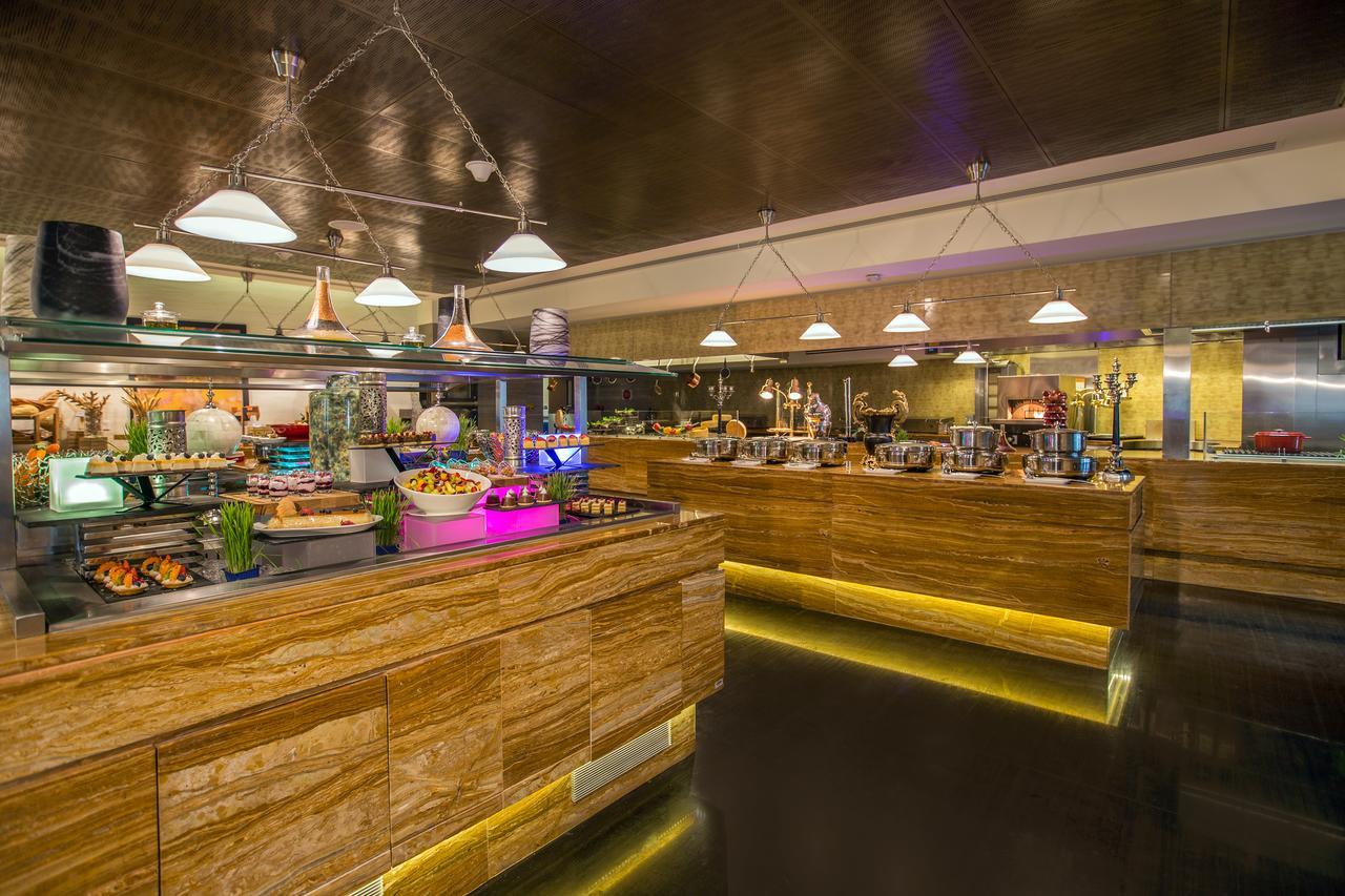 Hotel Millennium Plaza Doha Exterior foto The photo shows a modern buffet restaurant. There are several food stations with a variety of dishes displayed. The decor features wooden elements and soft lighting, creating a warm atmosphere. The counters are illuminated with subtle lighting, highl