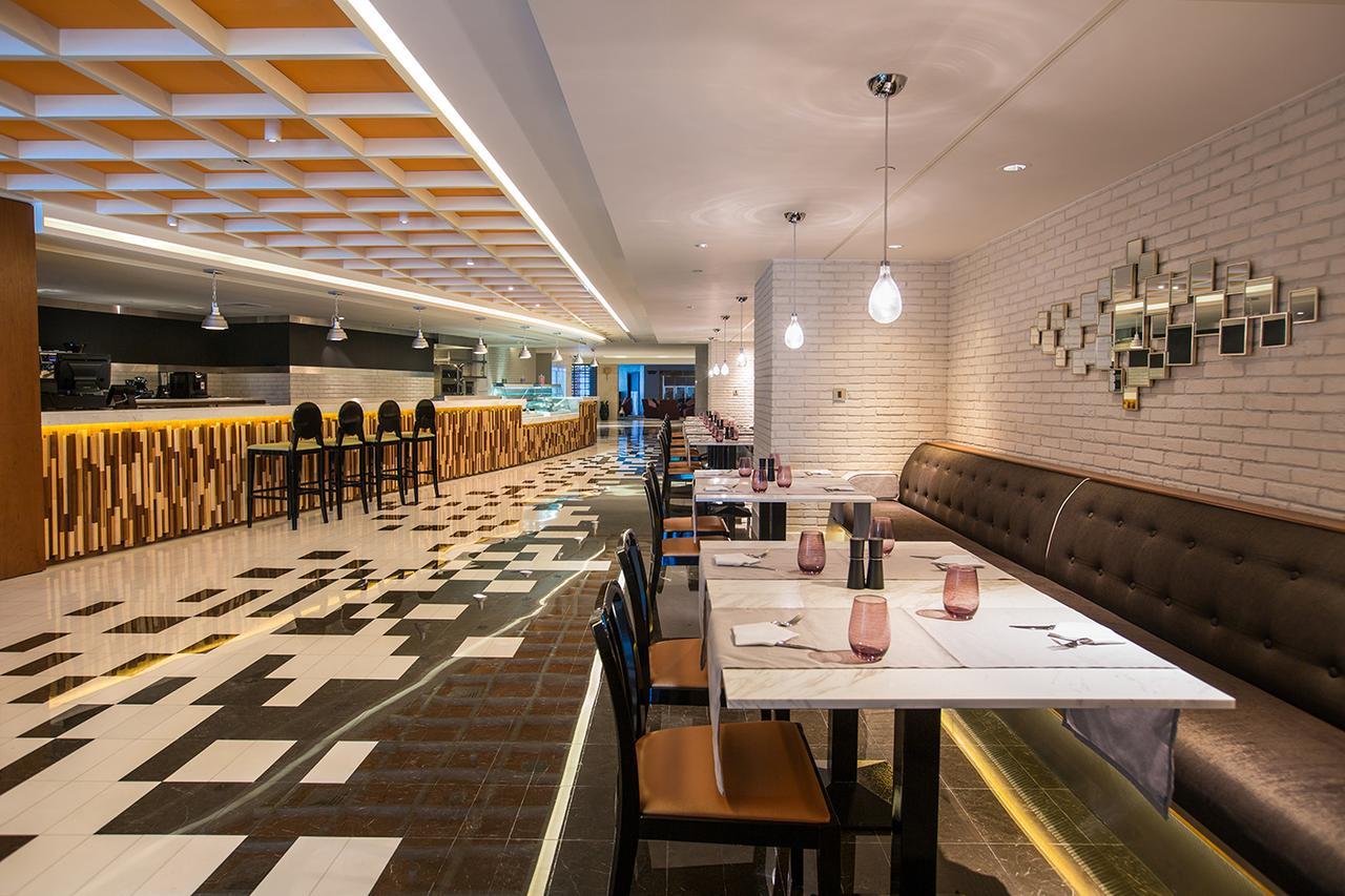 Hotel Millennium Plaza Doha Exterior foto The photo shows a modern restaurant interior. It features a long dining area with a combination of tables and a banquette. The flooring has a distinctive pattern, mixing black and white tiles. In the background, there's a bar area with high stools an