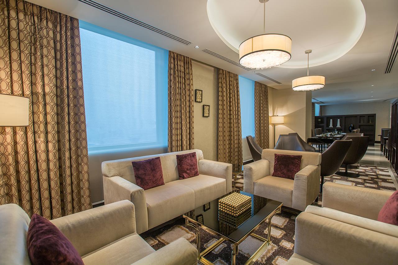 Hotel Millennium Plaza Doha Exterior foto The photo shows a modern living area characterized by a contemporary design. It features two light-colored sofas with purple cushions, arranged around a glass coffee table. The space is well-lit, with large windows covered by elegant curtains. There 