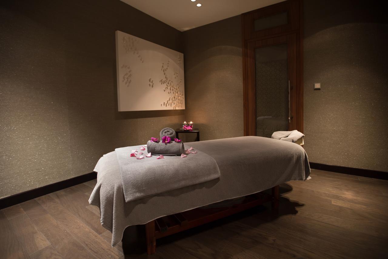 Hotel Millennium Plaza Doha Exterior foto The photo depicts a serene spa room designed for relaxation and massage therapy. The focal point is a massage table covered with a soft gray blanket, adorned with rolled towels and scattered flower petals. The walls have a muted, textured finish, and