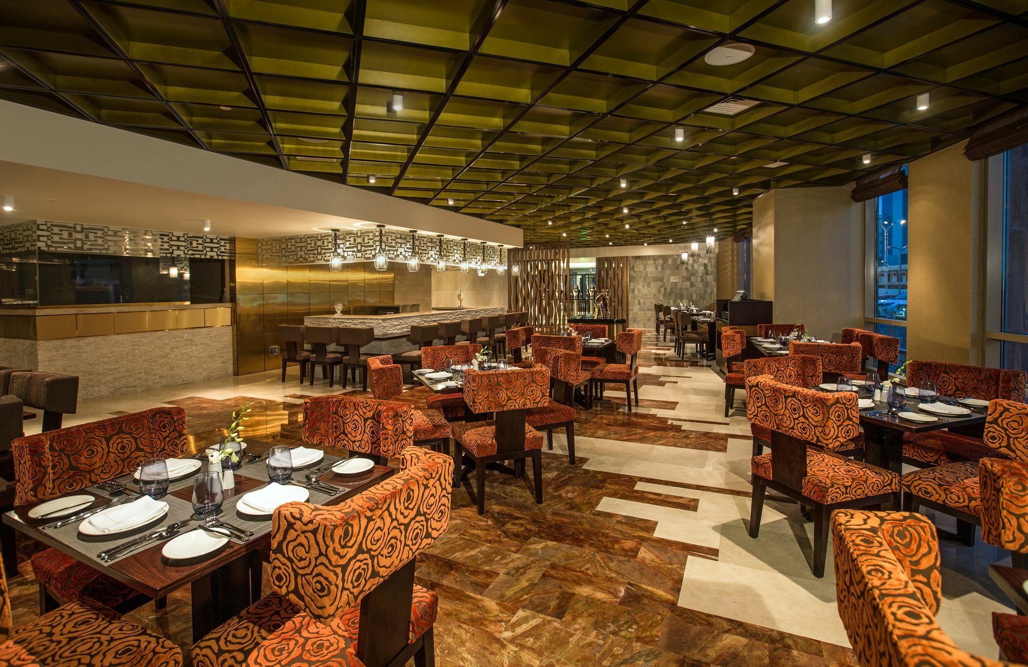 Hotel Millennium Plaza Doha Exterior foto The image shows the interior of a modern restaurant. The space features a stylish design with a green and brown color palette. Comfortable seating arrangements with orange patterned chairs surround tables set with plates and cutlery. The ceiling is d